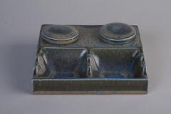 A pottery inkwell of dark blue color has a rectangluar shape body. There are two lid covering the inkwell and there are two pen holder.