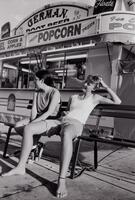 Two girls seated on a bench, one without shoes. A carnival food truck is behind them advertising popcorn.