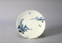 A medium size plate has blue-and-white underglaze painting of landscape. There are two groups of mountains; one of the left has two large rocks flanked by two smaller mountains with a few trees on the tops. Three smaller rocks peek from the water. On the right side, a range of gently sloped mountains and a rock are drawn. There are two fishing boats with masts can be seen in front of the mountain range. The crescent moon, shadowed by one side, is in the sky. The drawing is executed in thick underglaze and painted in lighter glaze. There are brown speckles on the surface; reddish color stains on the top of the rim. Some chips on the rim and on the back. The foot is unglazed; eye is glazed. There is a single band around the rim.