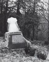 A woman in a white dress is walking away in a graveyard. There is a single headstone that is broken, trees behind.