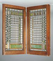 One of a pair of windows with a highly regular, rectilinear, although asymetric, design in both clear and colored glass.  The window has an oak frame.  Its design consists of vertical and horizontal bands of green and amber colorerd glass at the top and bottom of window; along one side are colored squares of glass, and along the other is a chevron-shaped column of glass. The overall effect is of colored pieces of glass suspended within a clear window, subdivided by abstract bands and patterns of lead caming.