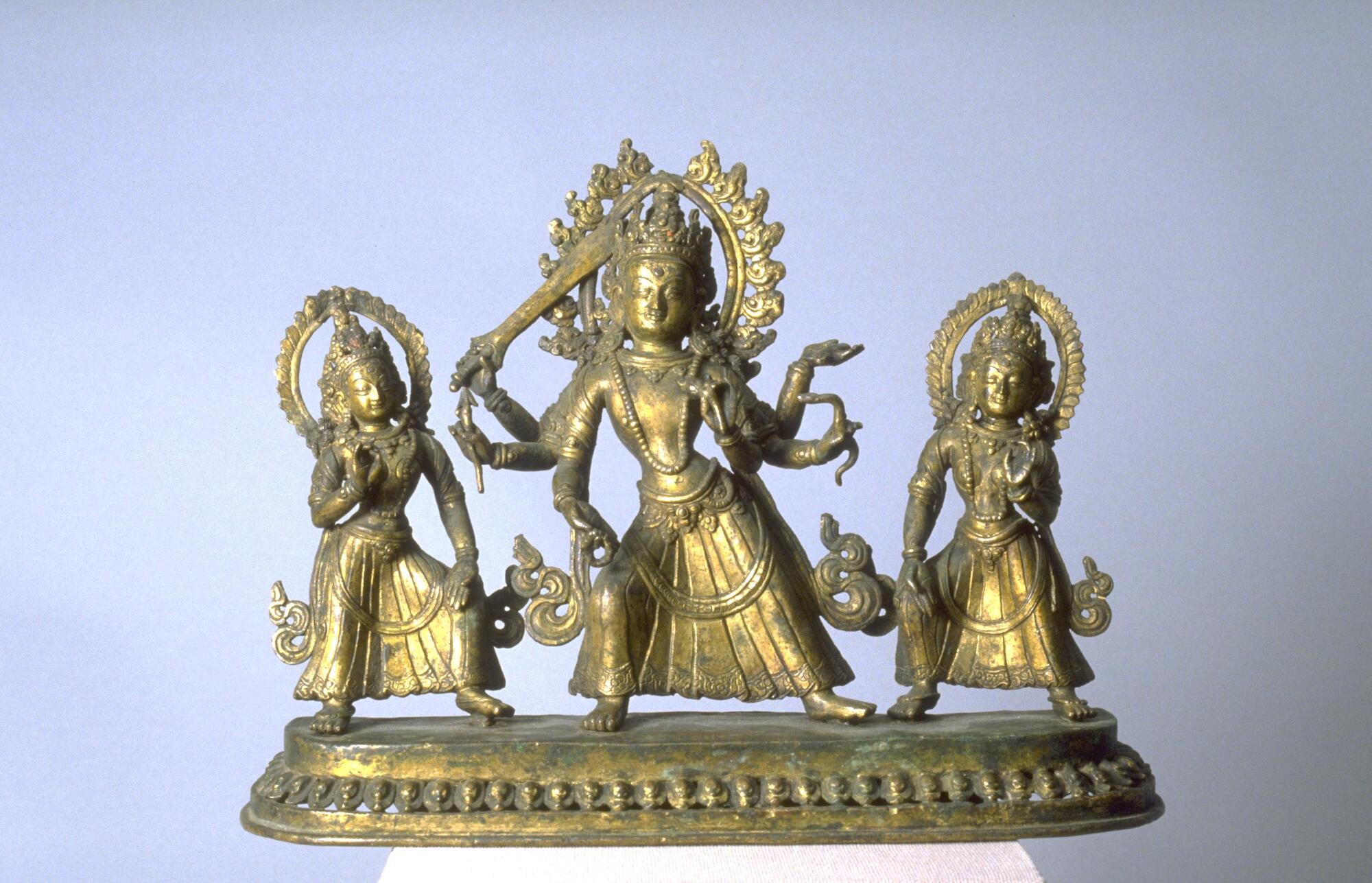 Mañjusri, the Bodhisattva of Wisdom, standing with two celestial attendants.  This representation of Manjushri includes six arms, one of which holds a sword, while a narrow book (modeled after books made from palm leaves) lays across his upper hand.  Manjushri is wearing an ornamented crown and necklace, and is encircled by a halo of flames.
