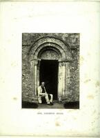 This photograph depicts a man sitting in the doorway of a church. He is framed by an arched stone carving. 