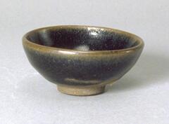 This round base bowl of light grey stoneware rests on a tall, straight foot ring. It is covered in a dark brown-black glaze with subtle hare's fur or <em>tuhao zhan (兔毫盏)</em> markings. 