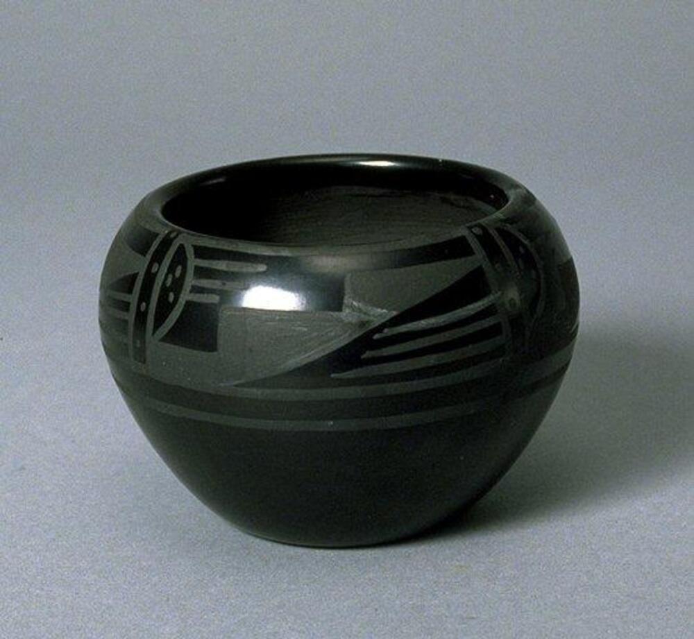 A deep bowl with a wide mouth made of black earthenware. The upper half of the bowl is decorated with a horizontal and diagonal design in a lighter shade of black and rougher texture than the smooth black surface.