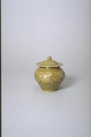 This is a squat stoneware jar with a wide globular shoulder on a narrow foot ring. Lotus petal lappets surround the base, and a peony scroll relief surrounds the body. The jar has a domed lid top with a wide flange and a ball knob; the area between is incised with scrolling meander. The object is covered in an olive green celadon glaze.