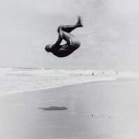 This photograph depicts a man mid-backflip on an ocean beach.
