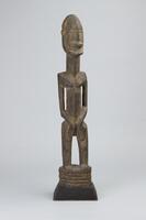 Standing male figure atop a ridged pedestal, all carved from the same piece of wood. The work is mounted on a newer wood base.&nbsp;