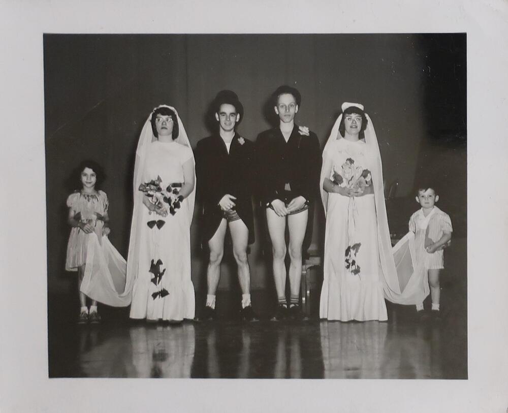 At the center of the image there are two men wearing long tuxedo jackets, top hats, and no pants. On the left and right of them are two women wearing light dresses with long veils. The women are holding bouquets and each has a small child standing behind her holding the veil train.