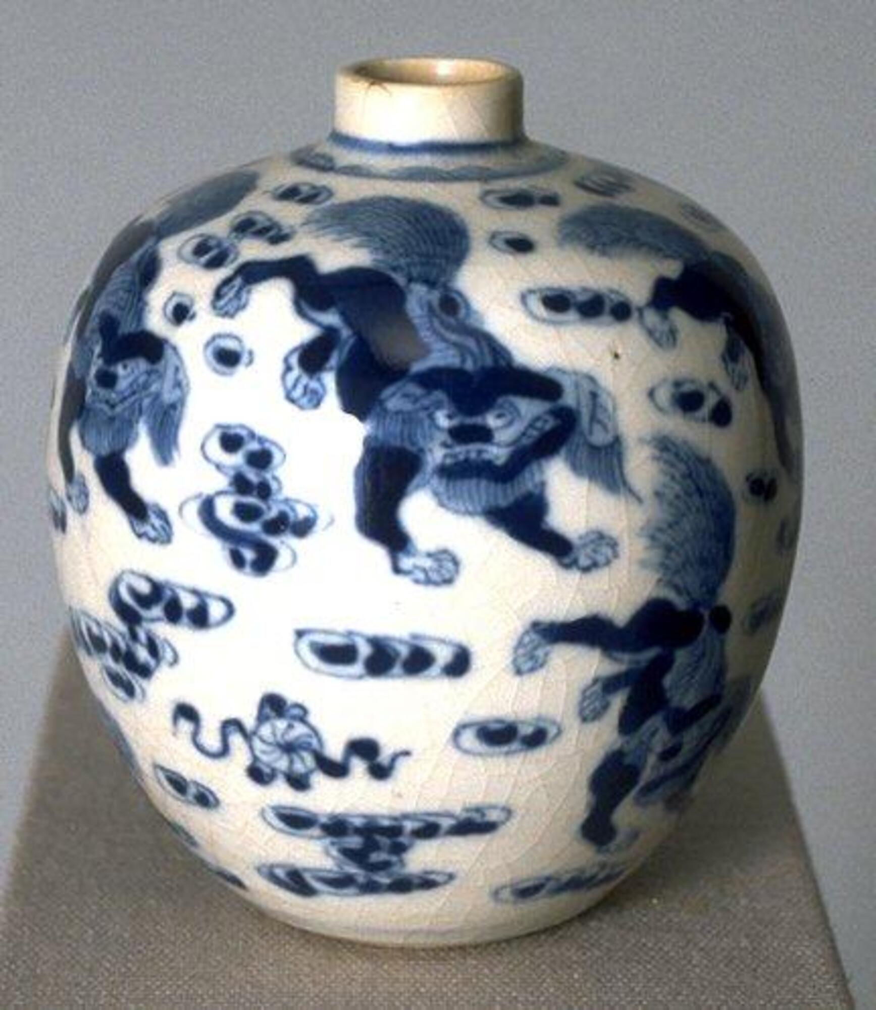 A porcelain globular jar with narrow straight neck and direct rim, on a recessed footring, covered in underglaze cobalt blue paintings of lions among clouds, then covered in clear glaze. It is missing the dome cap lid, and forms a pair with 1977/2.11.