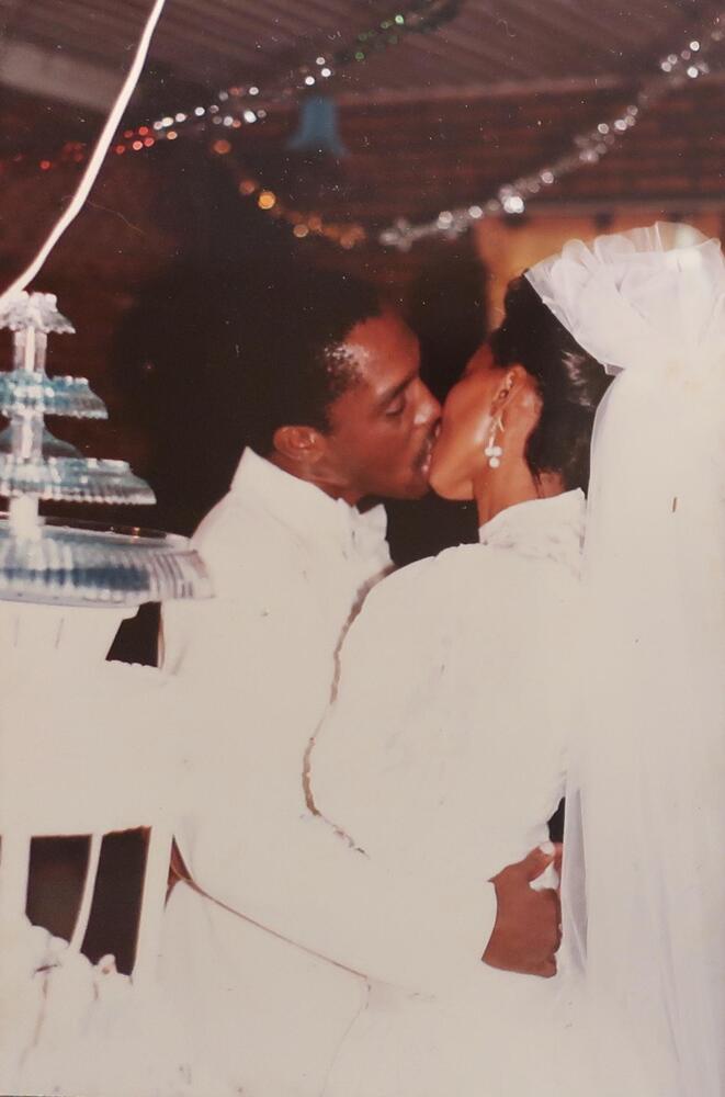 A couple kissing at what appears to be a wedding celebration. Both of them are dressed in all white and the woman wears a veil down her back. There are tinsel streamers hanging from the ceiling in the background.