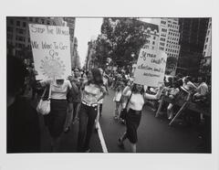 Photograph of a group of women protestors, smiling and holding posters, as they walk down a street fenced off to cars.