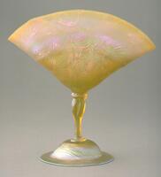 This vessel is in the shap of a fan, raised on a mold-blown stem, on a domed foot. The vessel has a raised design on both the fan and foot and is made from orange/pink iridescent glass.