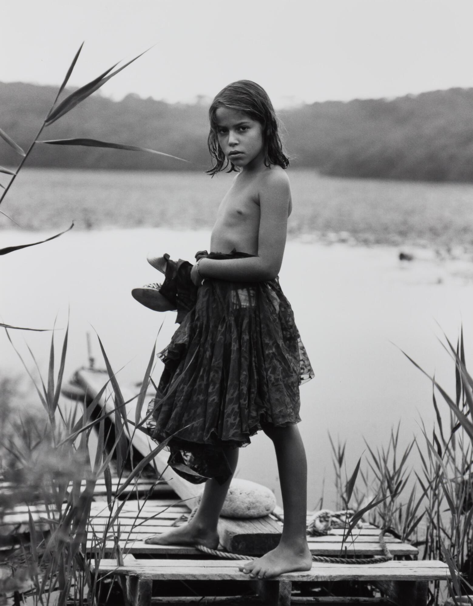 A child in a skirt, standing on a dock holding shoes.