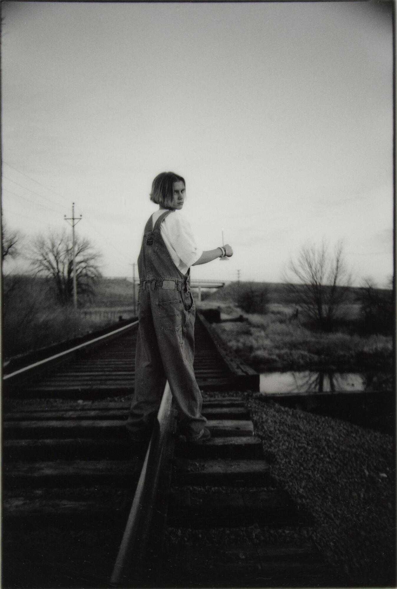 A girl in overalls standing on train tracks. She is looking back at the camera.
