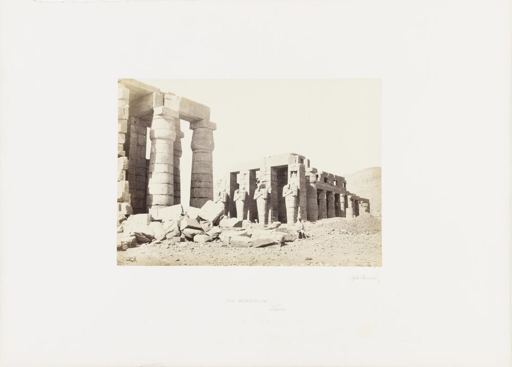 Two Egyptian buildings, each with columns, are depicted in a desert landscape with a man sitting on a pile of rubble in front of the complex.