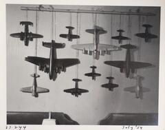 A hanging display of what appear to be model or toy airplanes. The planes are various sizes and are hung by their tales from a piece of bamboo mounted on the wall.