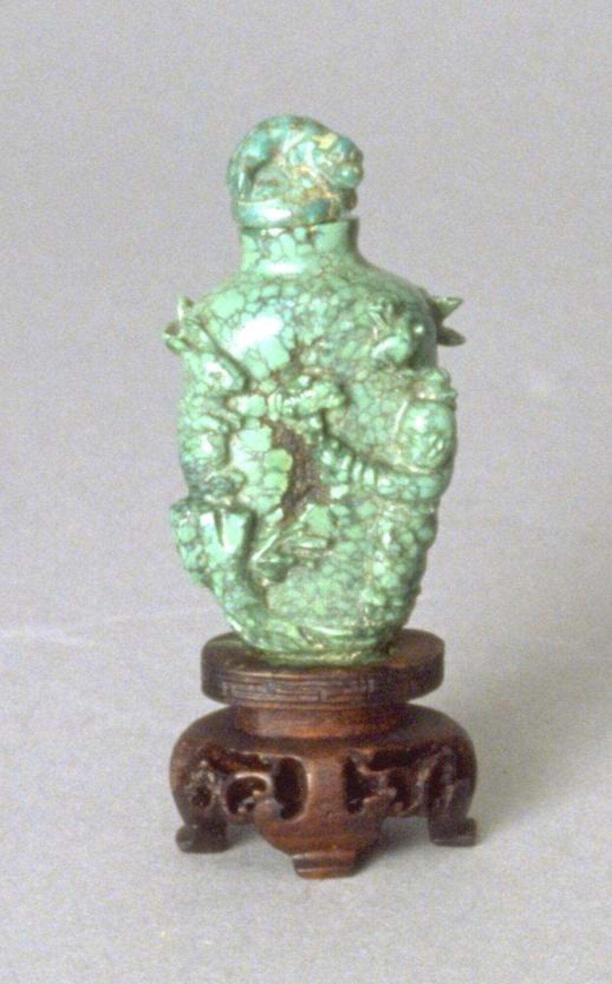 A crackle turquoise snuff bottle in the shape of a vase. Carved in high relief on the surface of the snuff bottle are rabbits and people. On the top of the snuff bottle is a crackle turquoise stopper. The snuff bottle is sitting on an incised wooden stand.