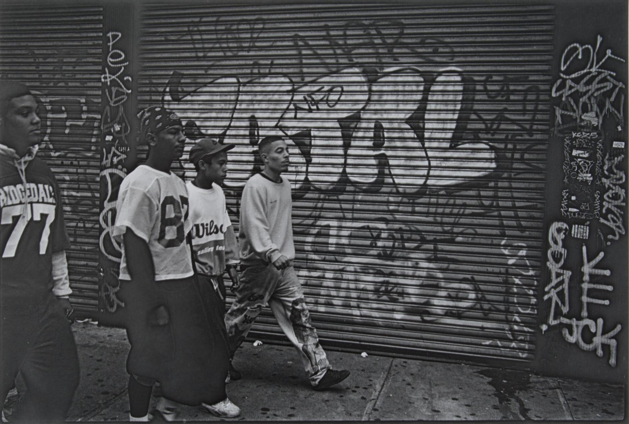 A photograph of a group of four young men in football jerseys and casual attire walking down the street. Behind them are two steel rolling doors covered in graffiti.