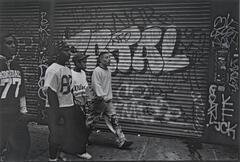 A photograph of a group of four young men in football jerseys and casual attire walking down the street. Behind them are two steel rolling doors covered in graffiti.