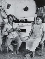 A man and woman sitting in wooden chairs in a kitchen with a white stove and stacked Corona boxes. He is barefoot and she is wearing sandasls. There are pots hanging on the walls.