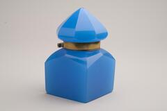 This is a bright blue glass inkwell with a brass collar. The body is square shaped with slanted shoulders. The blue glass lid is cone shaped with faceted sides.<br />