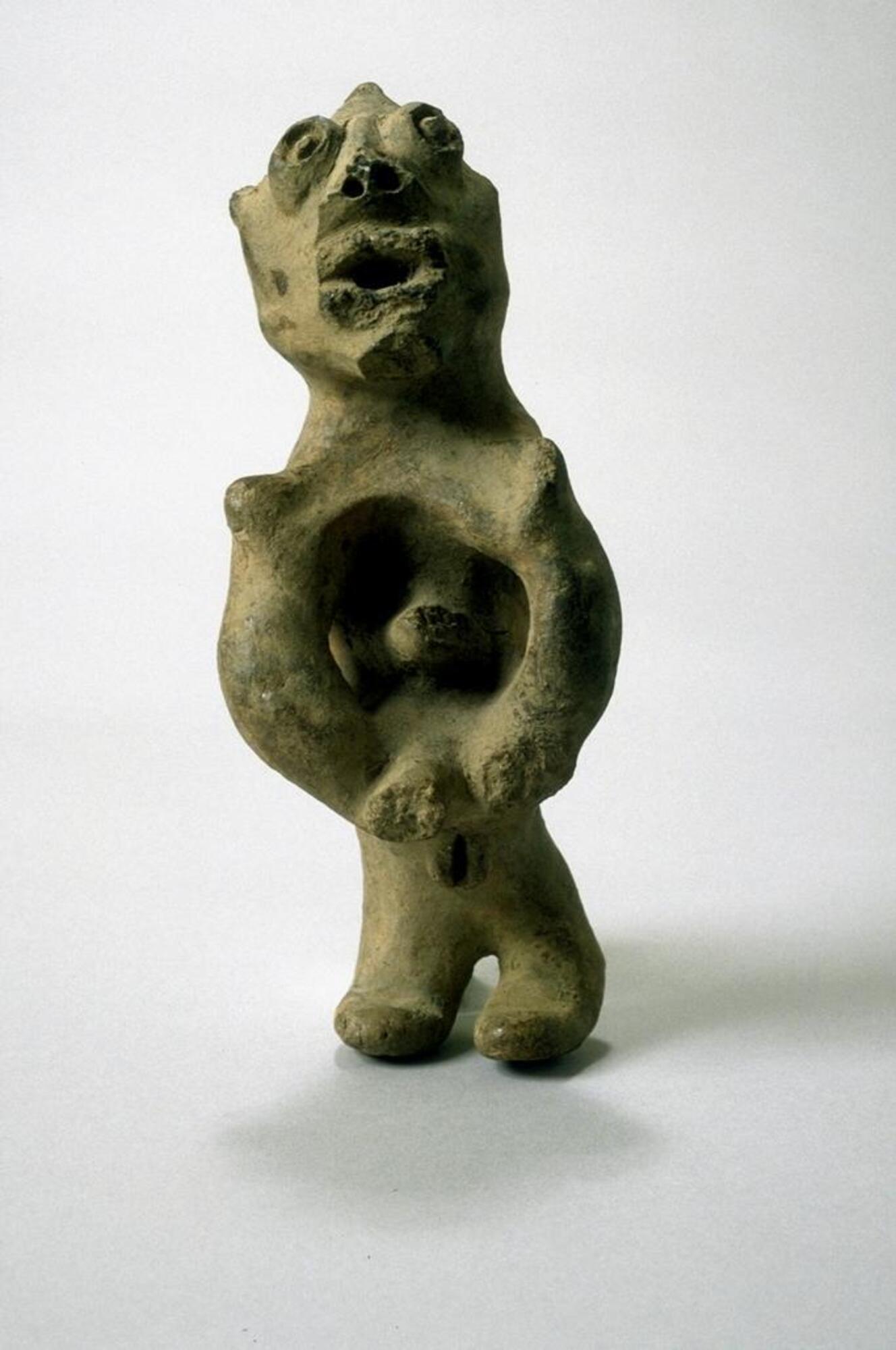 Standing female figure with short legs and a long torso. The arms are in front of the body and curve downward. Underneath the figure's hands is a cowrie shell embedded in the clay. The face of the figure looks upward with an open mouth and eyes formed by two concentric circles. 