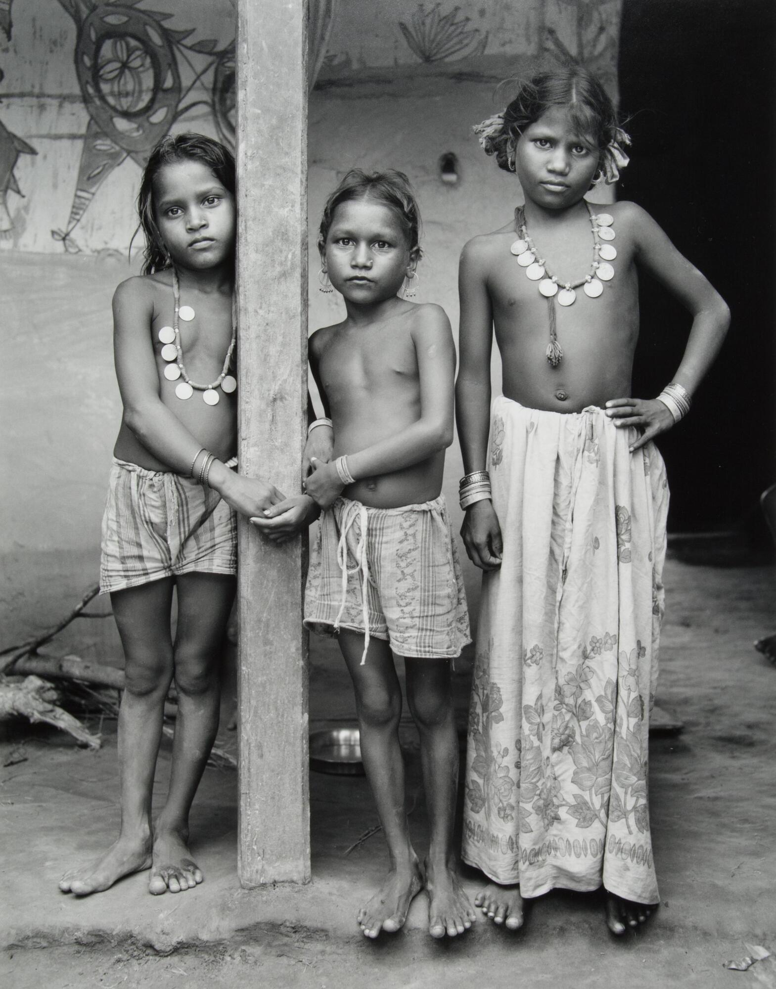 Three children standing next to a column. They are all barefoot and wearing similar beaded necklaces. Two are in shorts, one is in a long skirt.