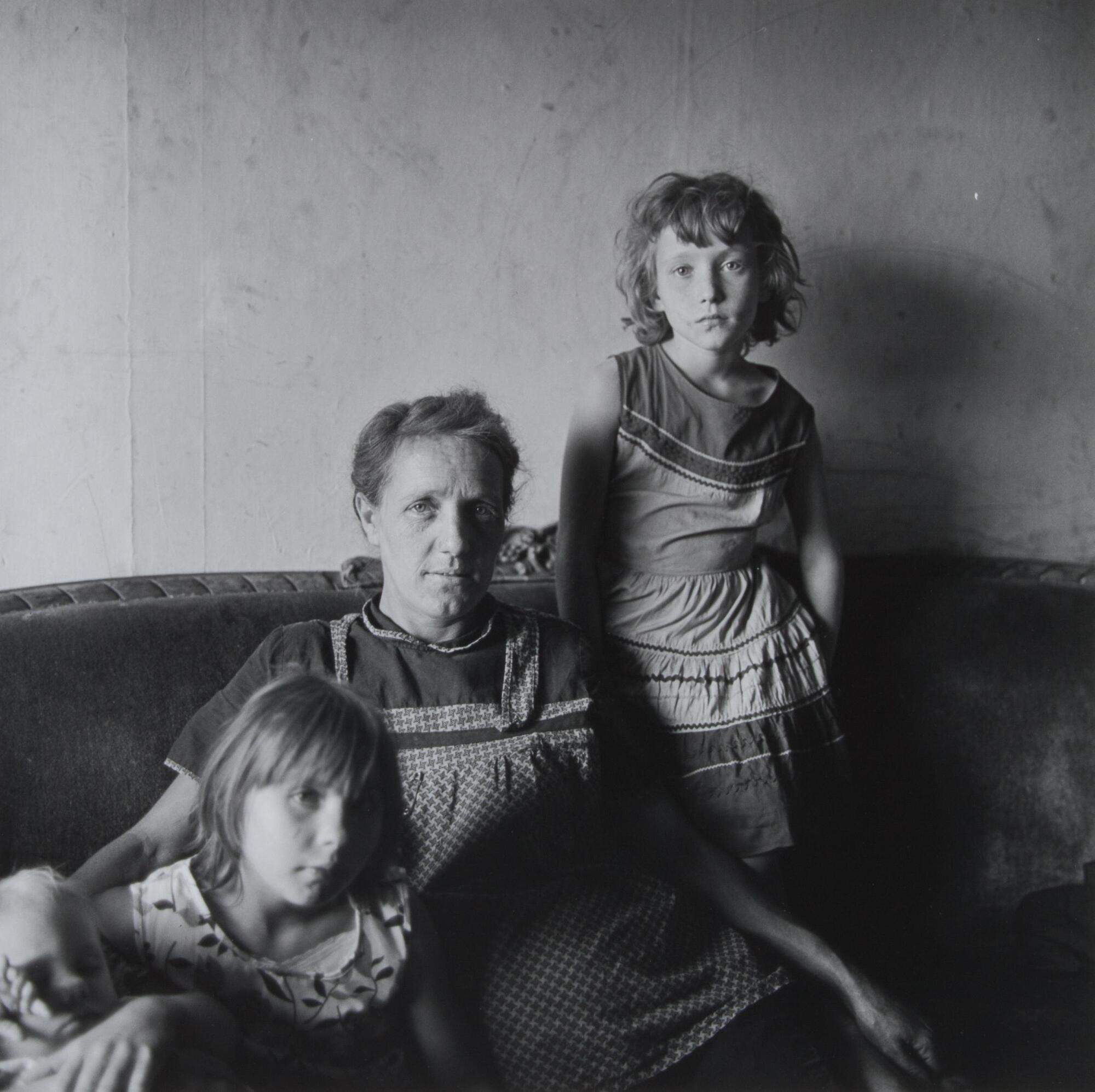 Portrait of a woman, her two daughters and a young baby. They sit on a sofa indoors; one daughter slouches into the couch on the left, holding the baby. The mother sits in the center, and the other daughter stands on the sofa cushion to the right of her mother.