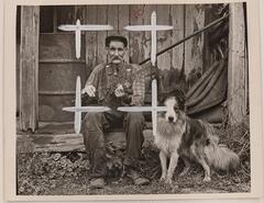 A man holding a violin sits on the porch of a wooden structure. A dog stands to the right of the man. Four white Xs drawn on the photograph outline a square around the man.&nbsp;