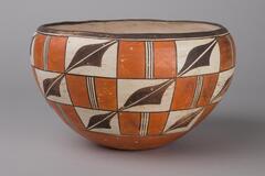 A squat, gracefully rounded pot with a mouth that is slightly smaller than the widest circumference of the pot, and a tapered base. The interior is colored white (slip). There is a band of black along the rim. The exterior is decorated with a checkerboard-like pattern of alternating rectangles of two designs: brownish-orange divided vertically by a white stripe broken by three thin black lines and white squares divided diagonally with wave-like black shapes.