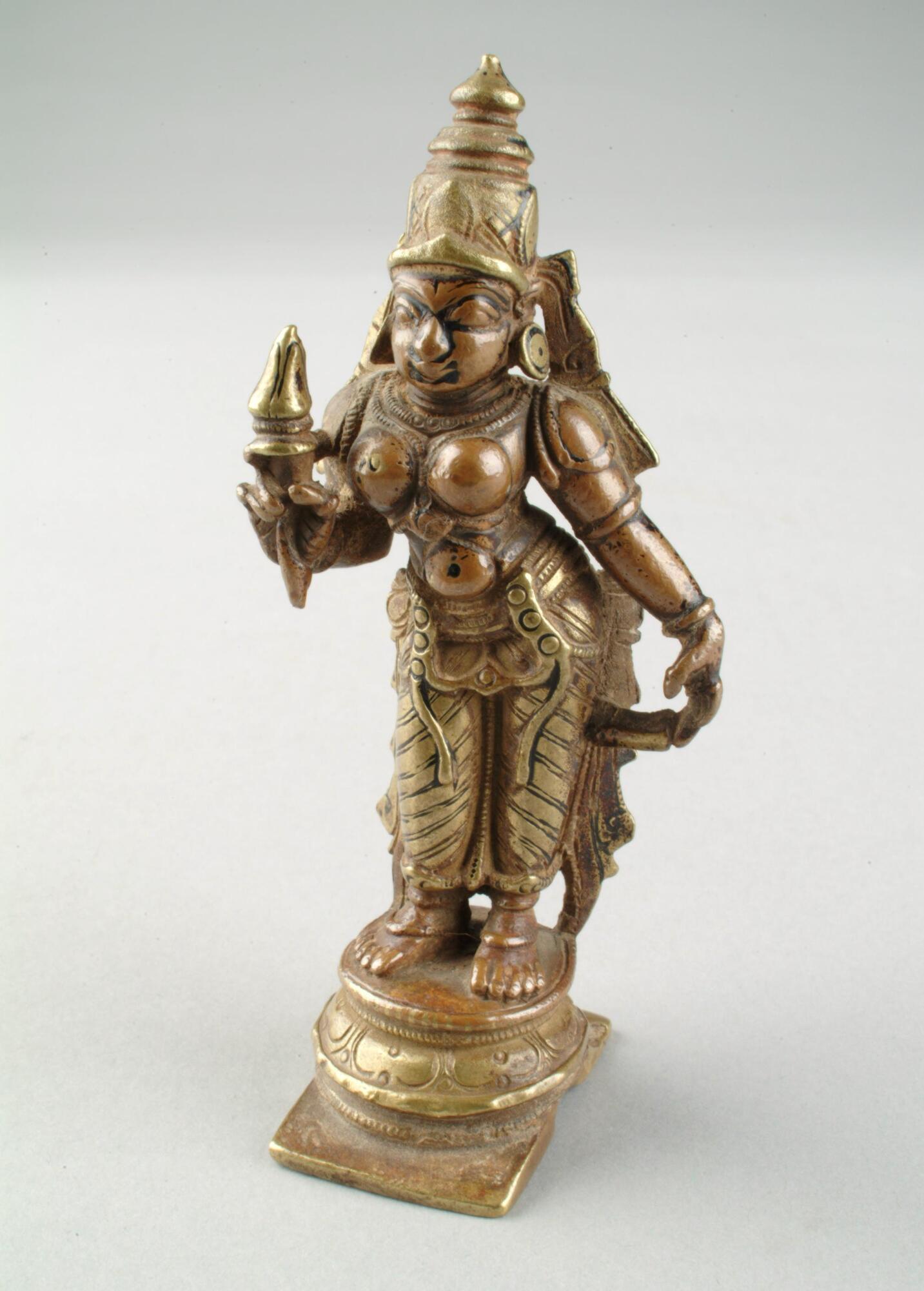 Bhudevi stands in a tribhanga pose (with three bends) with her left arm hanging pendant to her side and holding a lotus bud in her right hand. She leans towards the figure of Vishnu in the grouping of three bronzes.   She stands on a base consisting of a flat square element topped with a series of five round rings.  She wears a decorated lower garment flared out on either side in a pattern.  She wears a decorated belt and necklaces, bracelets and armlets, with shoulder loops, earrings and a crown.  The jewelry and crown is highlighted with gold paint as is his clothing and the two attributes.  She does not wear a band across her breasts, as does Shridevi in groupings with Vishnu and Bhudevi.<br />
