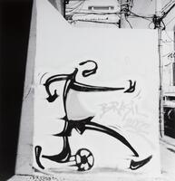 This photograph depicts a graffitied wall in an urban environment. Spray painted on the white wall is a stylized running figure playing with a soccer ball, with the words “Brasil 2002” written next to it.<br />
 