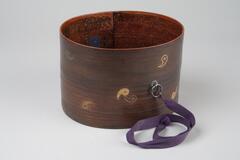 Oval wooden container with circular designs around. There are metal hooks and purple ribbons. This is a part of a portable tea set.