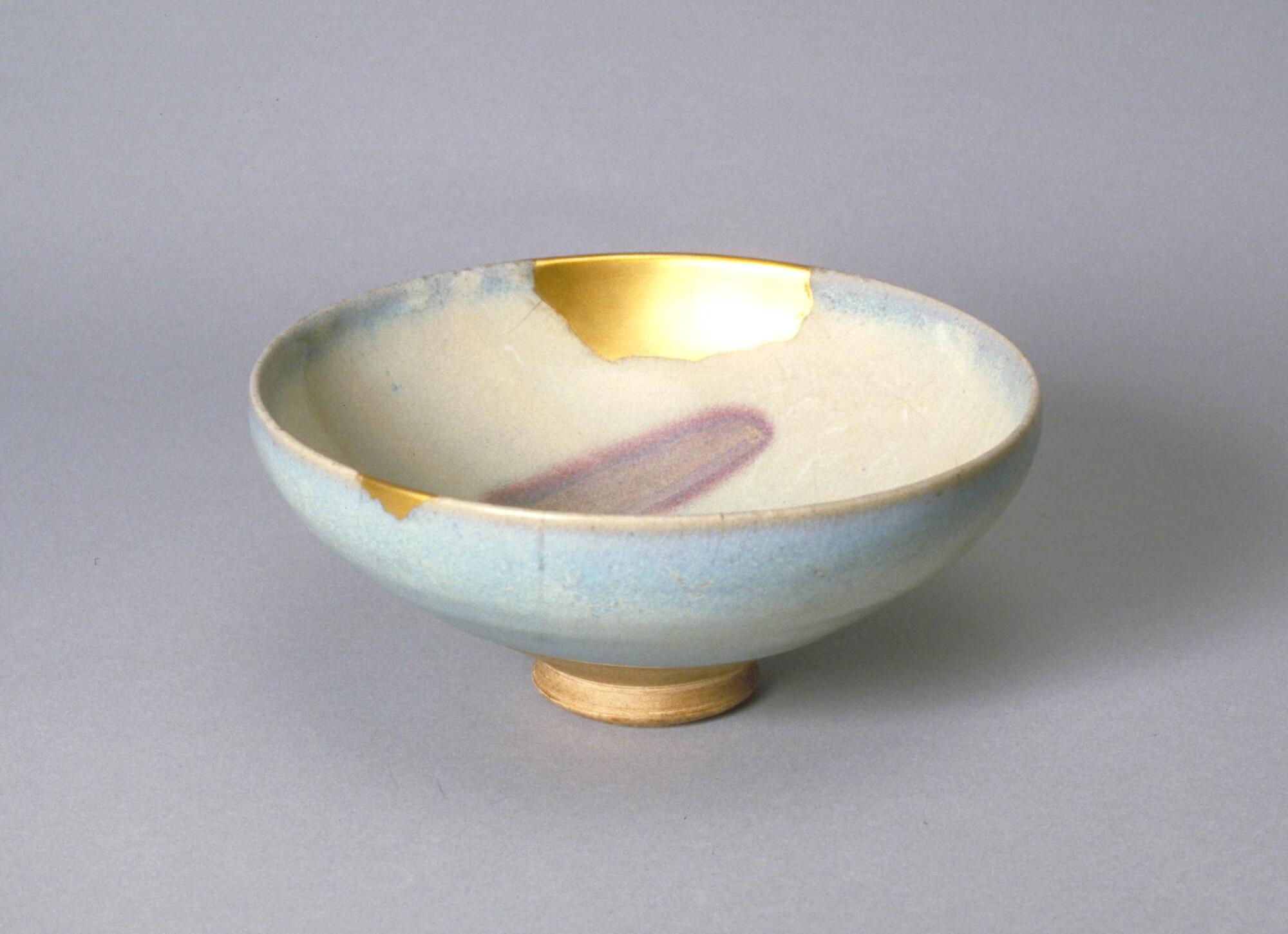 This shallow conical bowl on a straight footring is covered in a pale blue opaque glaze, the interior is moon-white and the exterior, sky blue. There is a copper reddish-purple splash to the interior and two gold repairs along the rim and side.
