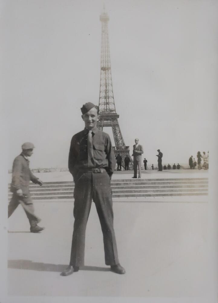 A young man in military uniform standing in front of the Eiffel Tower. He poses with his feet apart and his hands behind his back, smiling at the camera.