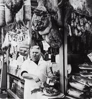 A man stands in the threshold of a shop with meats and cheeses on display all around him.