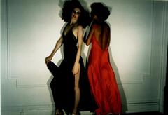 A color photograph of two models standing in the center of the frame. The model to the left wears a dark navy satin nightgown. She turns her body toward the left side of the frame, gathering the gown in her right hand. The model to the right wears a bright red gown and faces the wall, turning to the side as if to tell a secret.