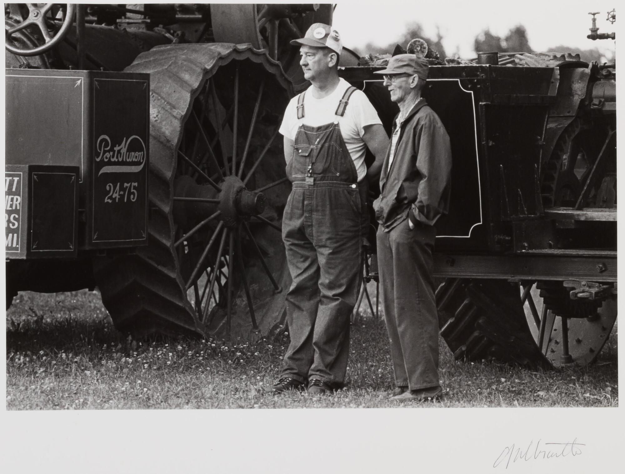 Two farmers standing next to a large tractor, one in overalls with his hands behind his back, the other in a dark jacket with his hands in his pockets.