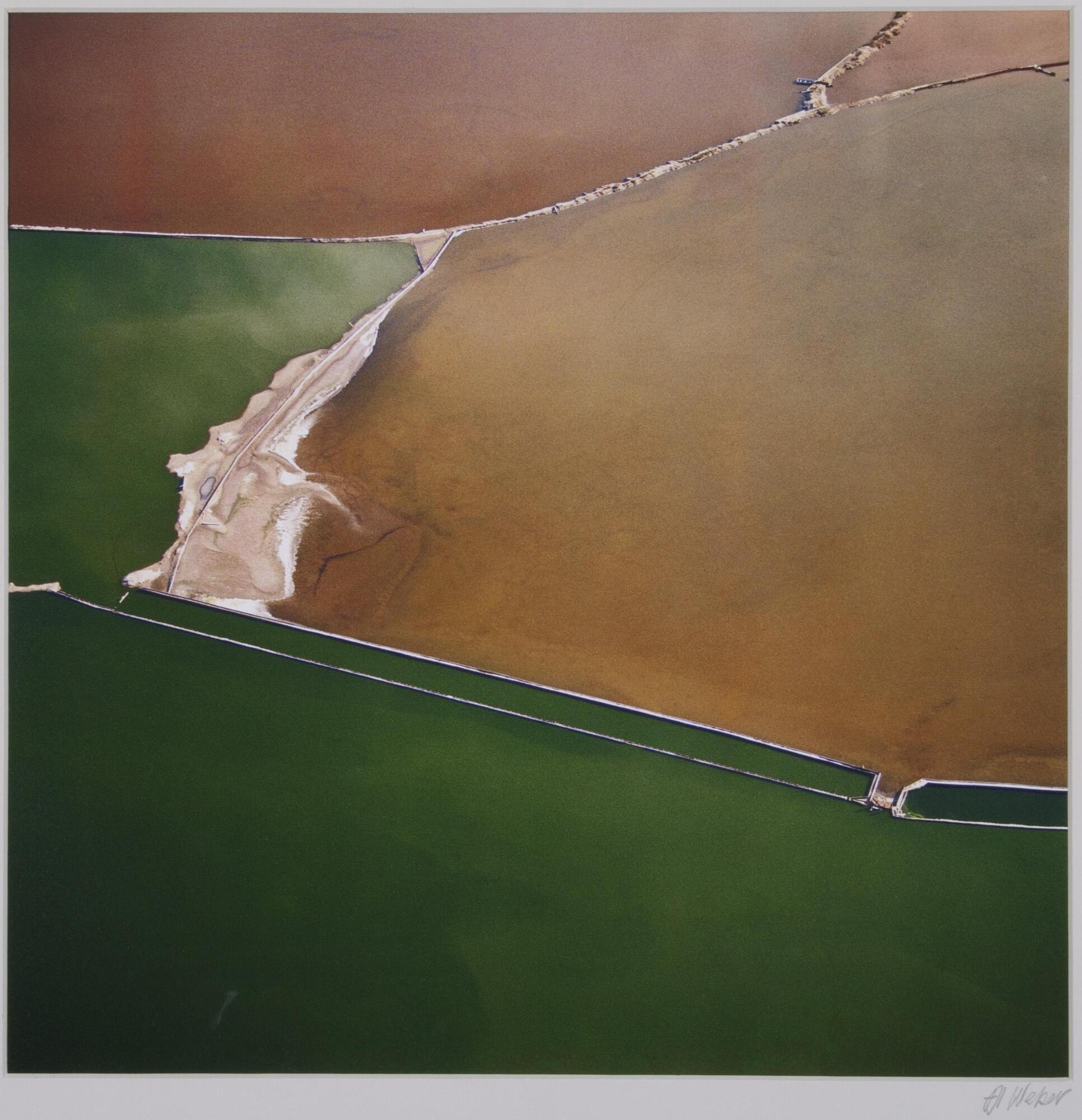 An aerial view of land meeting water. The water is a deep emerald green and the land, a varying shade of ochre and brown. The lower section is the water and there is a boat dock on the lower right section of the image. The land mass is the upper portion of the image, but seems to be separated at one section in a horizontal format.