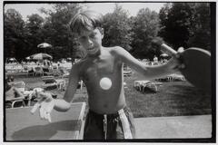 A portrait of a young boy playing ping pong, the ball suspended in mid-air between his hands. Behind him, a lawn with lounge chairs and a pool are visible.