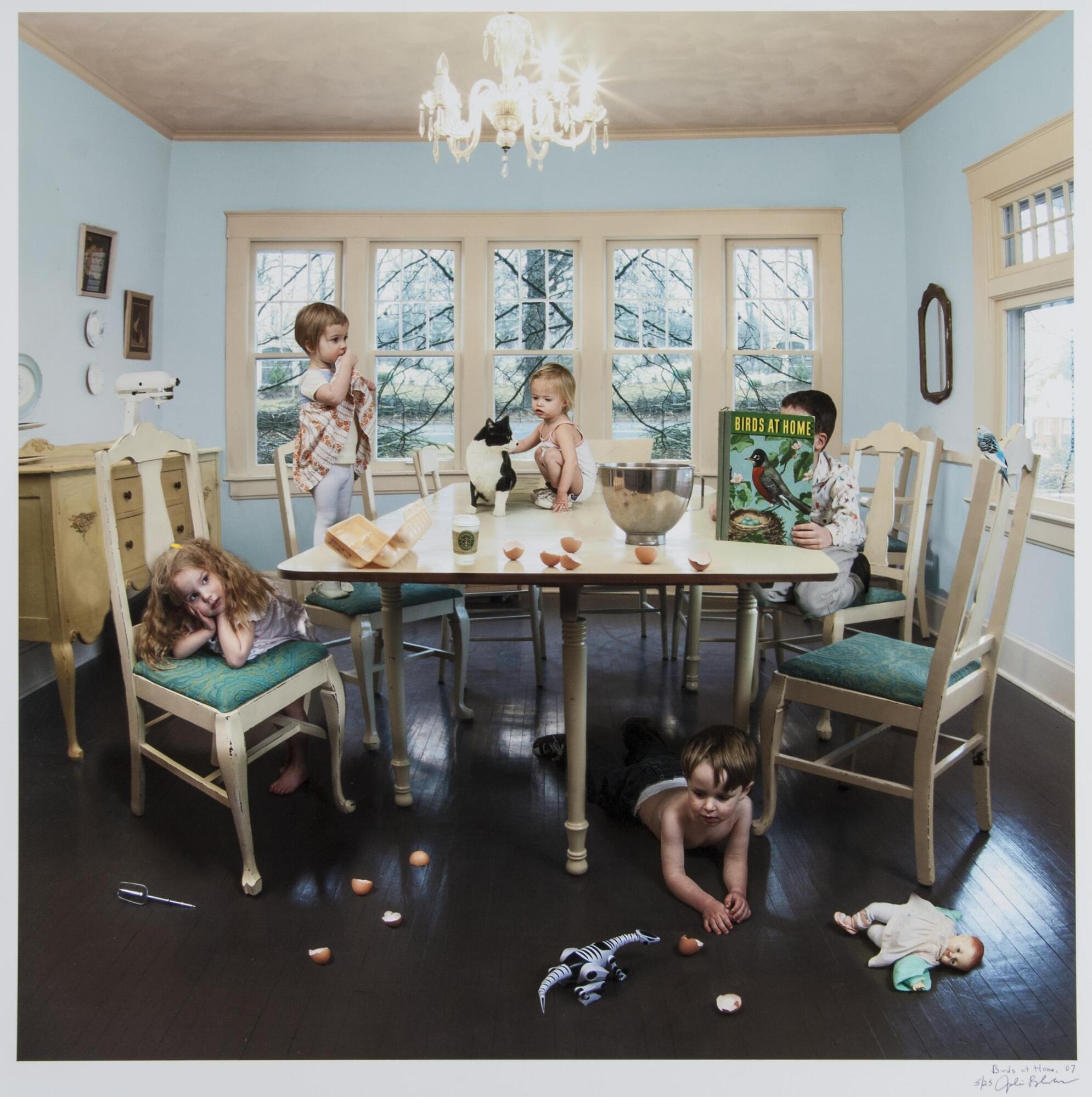 A color photograph of a group of children and a cat at a table. Each child engages with something in the room, which is in an organized disarray. Egg shells fall from the table to the floor, toys are strewn about, and an infant and cat sit in the middle of the table.