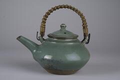 A wide diamond-angled teapot and lid with straw (or wood) wrapped handle. The pot is a blue-green color with copper-colored flecks throughout. Green glaze.