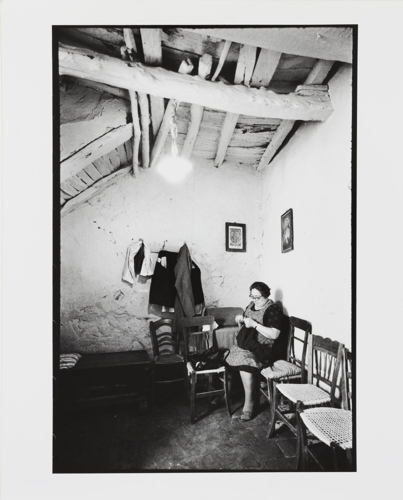 A woman sits in the corner of a room with white-washed walls and ceiling rafters. Chairs line the wall to her left.