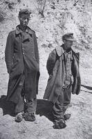 Two men in tattered winter clothing and cloth-wrapped feet stand amid rocky terrain. 
