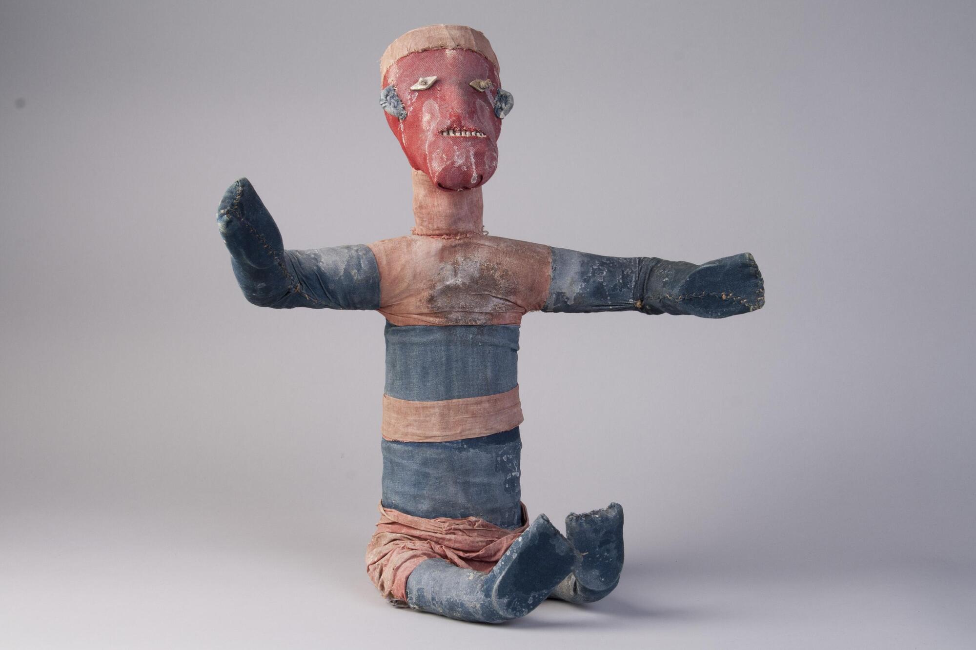 Cloth figure of a human, bound around human remains. The figure is in a seated position with arms outstretched. The torso and neck are cylindrical and elongated.