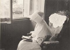 A nun seated on a rocking chair near a window reading a book.