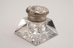 This is a clear cut glass inkwell with a sterling silver lid. The body has the shape of a cropped pyramid with triangle patterns on the edges and starburst designs. The silver lid has a round shape with floral designs.