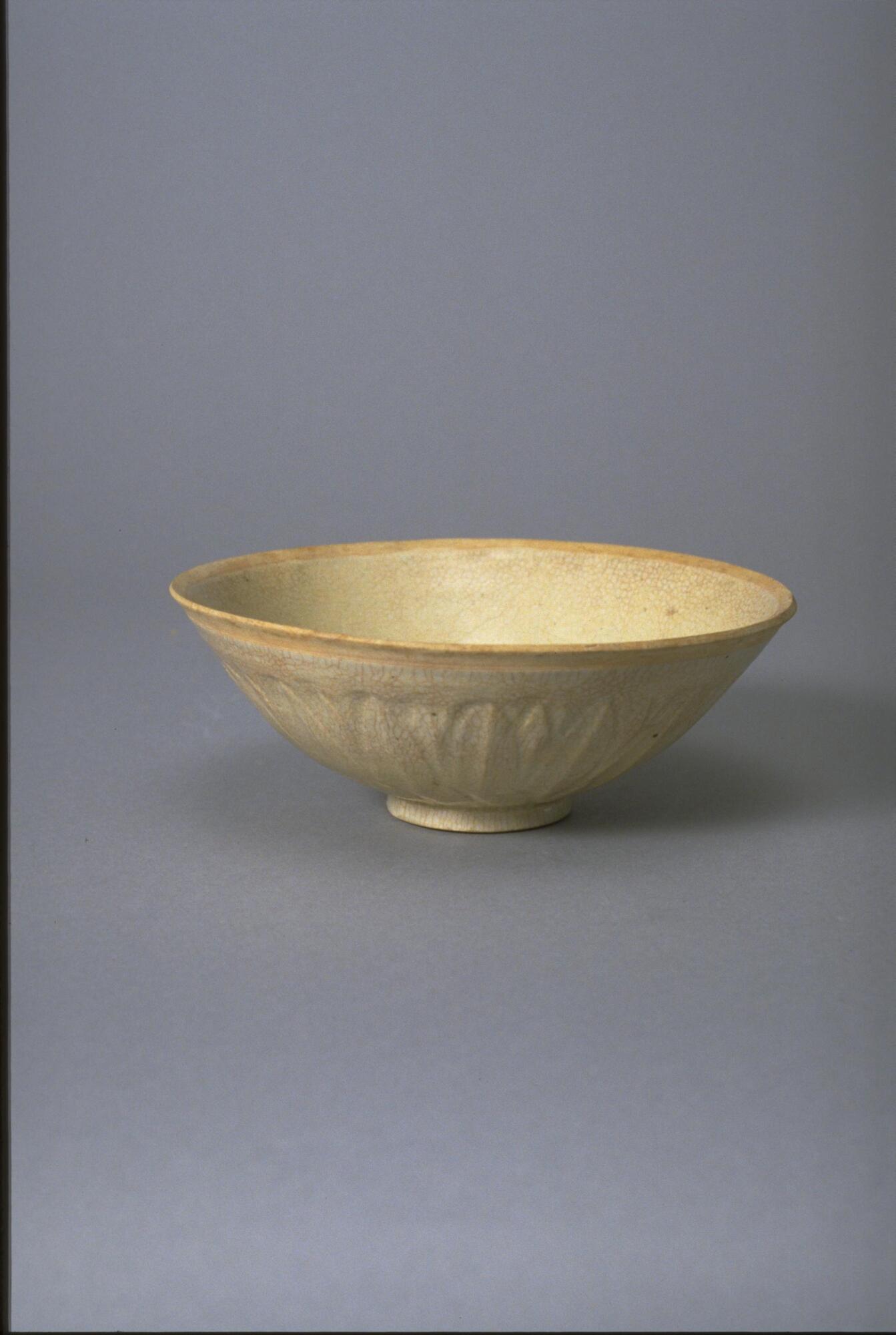 This thin porcelain conical bowl with direct rim on a footring has an exterior carved with lotus petal decoration.  It is covered in a white glaze with bluish tinge, and the rim is unglazed.