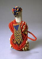 Wooden columnar figure heavily decorated with beads. The top of the figure has a cluster of thread, each with 2 or 3 beads attached. The neck of the figure is covered with white seed beads. Clusters of cowrie shells form the limbs of the figure. The body of the figure is covered with strands of red beads. One side of the figure has a blue and yellow chevron patterned beadwork with fringe.